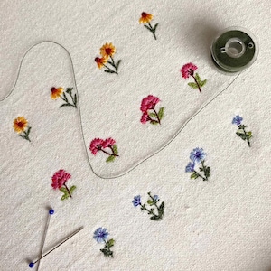 以下が含まれることがあります： 白い布に手刺繍された花の刺繍。 黄色、ピンク、青の色合いの小さなカラフルな花と緑の葉が特徴です。 緑色の糸巻きと2本のピンも表示されています。