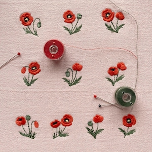 Peut inclure: Gros plan sur un tissu rose avec des coquelicots rouges brodés et des feuilles vertes. Deux bobines de fil, une rouge et une verte, sont présentes, ainsi que deux épingles. Les coquelicots sont en fleurs.