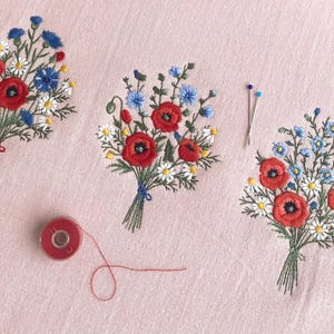Puede incluir: Bordados florales de ramos con amapolas rojas, acianos azules y margaritas blancas sobre tela rosa. También hay una bobina de hilo rojo y alfileres.