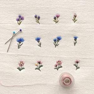 May include: Embroidered floral design on white fabric. The design features small, colorful flowers in shades of purple, blue, and pink. Two pins and a pink thread spool are also visible, suggesting a craft project.