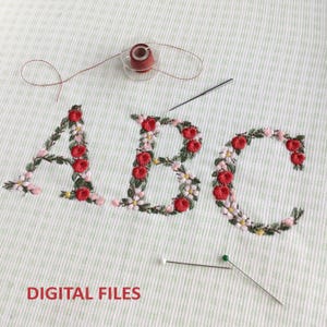 May include: Embroidered letters A, B, and C in a floral wreath design. The letters are stitched in red and white thread on a white fabric background. The letters are surrounded by green leaves and small red and white flowers.