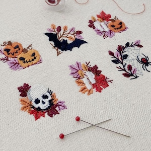 Pode incluir: Desenhos bordados com tema de Halloween, incluindo um morcego, abóboras, uma caveira, uma aranha e velas. Cada desenho é rodeado por folhas de outono em tons de laranja, vermelho e roxo. Bordado em tecido creme, com alfinetes e linha visíveis.
