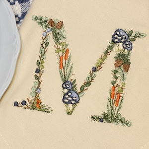 Op de afbeelding: Een beige linnen servet met een geborduurde letter "M" gemaakt van groene bladeren, blauwe paddenstoelen, oranje bessen en bruine dennenappels.