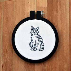 May include: A black and white embroidery of a cat sitting on a white fabric stretched over a black embroidery hoop.