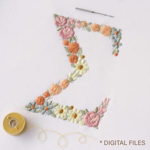 Op de afbeelding: De letter Z is gevormd door geborduurde bloemen in tinten roze, oranje, geel en saliegroen op witte stof. Een zilveren naald rust boven de letter en een kleine houten klos met geel garen ligt eronder, met de tekst DIGITAL FILES.