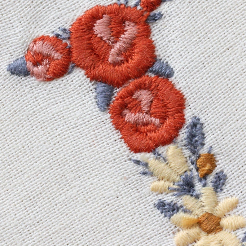 May include: Close-up of embroidered flowers in shades of orange, pink, and yellow on a white fabric background. The flowers are arranged in a vertical line, with the largest flower at the top and the smallest flower at the bottom.