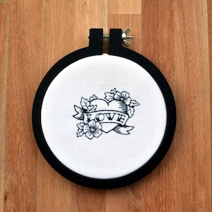 Puede incluir: Aro de bordado negro con tela blanca que presenta un corazón bordado en negro con la palabra "LOVE" en una pancarta y detalles florales.