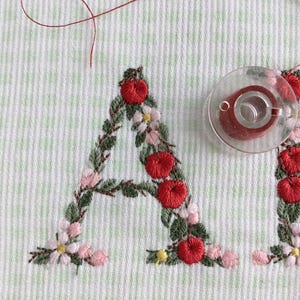Puede incluir: Primer plano de un dise&ntilde;o de bordado hecho a mano de la letra "A" con bayas rojas, hojas verdes y flores rosas. El bordado est&aacute; sobre una tela blanca con un sutil patr&oacute;n de cuadr&iacute;cula.