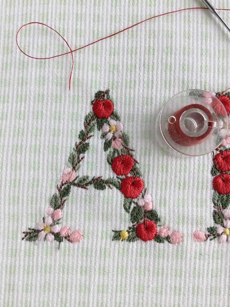 Puede incluir: Primer plano de un dise&ntilde;o de bordado hecho a mano de la letra "A" con bayas rojas, hojas verdes y flores rosas. El bordado est&aacute; sobre una tela blanca con un sutil patr&oacute;n de cuadr&iacute;cula.