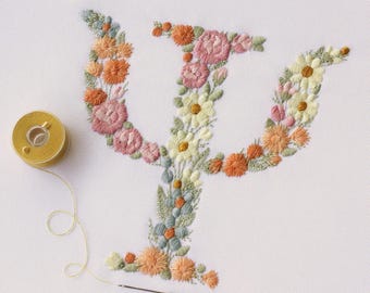 Puede incluir: Una superficie de tela blanca con la letra griega Psi bordada, compuesta por varios patrones florales en tonos rosa, naranja, amarillo y verde. A la izquierda hay un carrete de hilo dorado con una aguja e hilo. El texto dice: * DIGITAL FILES