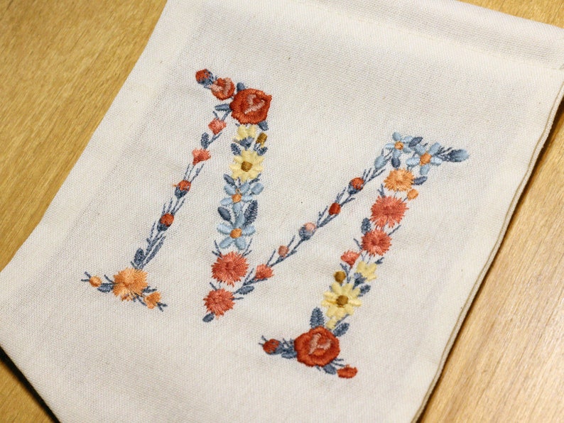 Pode incluir: Uma bolsa de lona branca com a letra M bordada em flores laranja, amarelo e azul.