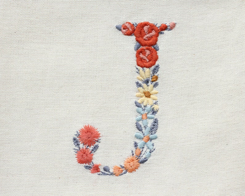 May include: Embroidered letter J made of colourful flowers on a white fabric background. The letter is formed by a variety of flowers in shades of red, pink, blue, and yellow.