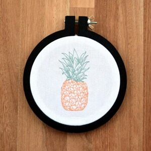 Puede incluir: Diseño de piña bordado en una tela blanca estirada en un aro de bordado negro.