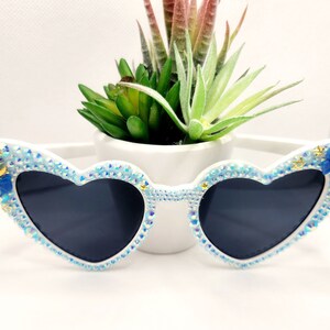 Può includere: Occhiali da sole bianchi a forma di cuore con decorazioni blu e dorate. Gli occhiali hanno un design a mezzaluna e stella su un lato e un fiocco blu sull'altro.
