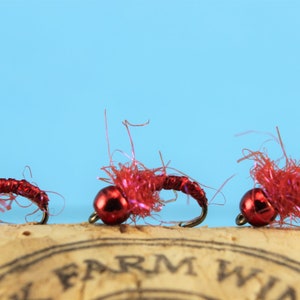 Puede incluir: Tres se&ntilde;uelos de pesca con mosca de cabeza de cuentas rojas con hilo de seda rojo y plateado. Los se&ntilde;uelos est&aacute;n sobre una superficie de corcho.