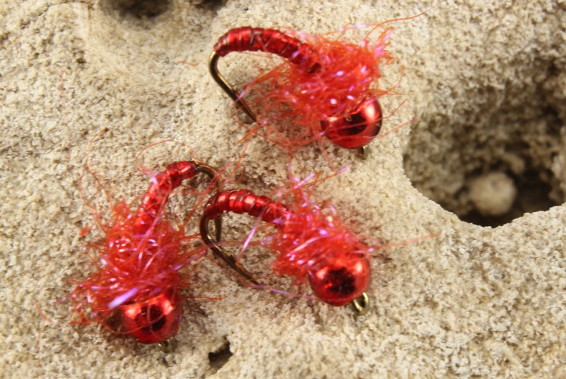 Puede incluir: Tres se&ntilde;uelos de pesca con mosca rojos con un acabado met&aacute;lico brillante y un cuerpo rojo peludo.