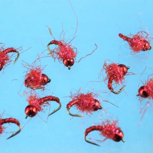 Puede incluir: Primer plano de diez se&ntilde;uelos de pesca con mosca rojos con anzuelos dorados y cuentas rojas. Los se&ntilde;uelos est&aacute;n esparcidos sobre un fondo azul.