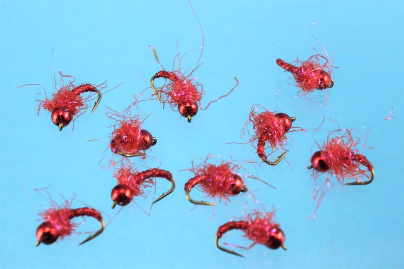 Puede incluir: Primer plano de diez se&ntilde;uelos de pesca con mosca rojos con anzuelos dorados y cuentas rojas. Los se&ntilde;uelos est&aacute;n esparcidos sobre un fondo azul.