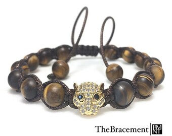May include: A brown beaded bracelet with a gold tiger head charm. The charm is encrusted with clear rhinestones and has black eyes. The bracelet is made with brown cord and tiger eye beads.