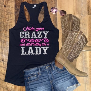 May include: Black tank top with the text "Hide your crazy and start acting like a lady" in silver glitter with pink accents. The tank top is paired with denim shorts and brown cowboy boots.