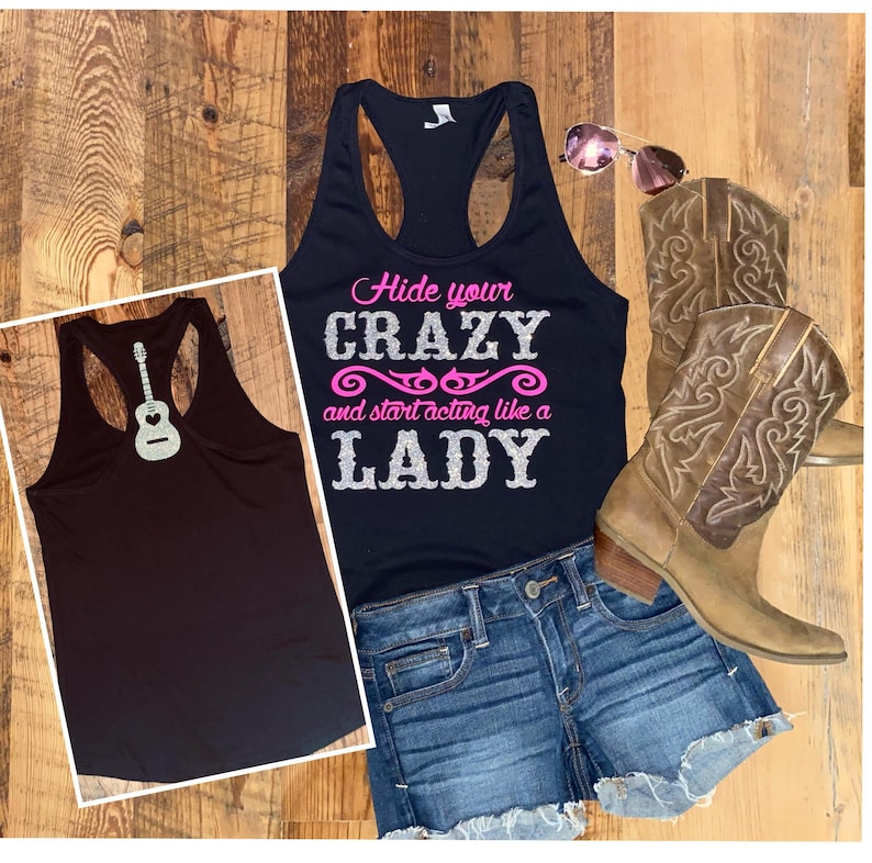 May include: Black tank top with the text "Hide your crazy and start acting like a lady" in pink and silver glitter. The tank top is shown on a model wearing denim shorts and brown cowboy boots.