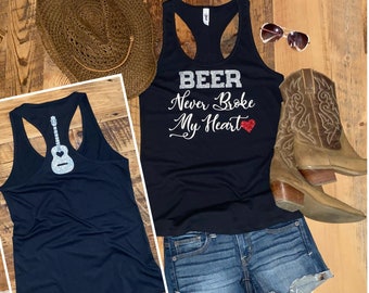 May include: A black tank top with the text "BEER Never Broke My Heart" in silver glitter and a red glitter heart. The image also includes a straw cowboy hat, denim shorts, sunglasses, and brown cowboy boots.