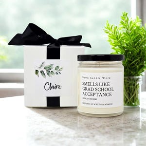 May include: A white gift box with a black ribbon and a label reading Claire, next to a glass jar candle with a black lid. The candle label reads Dawn Candle Worx, SMELLS LIKE GRAD SCHOOL ACCEPTANCE, NATURAL SOY WAX CANDLE, HAND POURED, NET WT 9OZ.