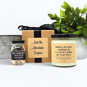 May include: A gift set with a candle, matches, and a box. The candle has a label that reads "Smells Like Aaron Is A Lucky Son Of A B*tch." The match jar is labeled "The Perfect Match." The box has a label that reads "And the Adventure Begins..."
