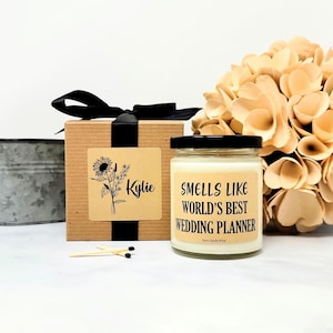 May include: A candle in a glass jar with a black lid, labeled "Smells Like World's Best Wedding Planner." A gift box with a black ribbon and a floral design is next to the candle. Matches and a bouquet of flowers are also in the image.