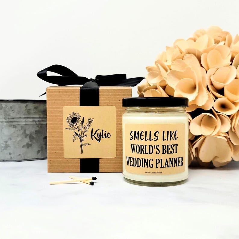 May include: A candle in a glass jar with a black lid, labeled "Smells Like World's Best Wedding Planner." A gift box with a black ribbon and a floral design is next to the candle. Matches and a bouquet of flowers are also in the image.