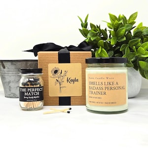 May include: A gift set featuring a candle, matches, and a decorative box. The candle has a label that reads "Smells Like a Badass Personal Trainer." The match jar is labeled "The Perfect Match." The box is adorned with a sunflower design and the name "Kayla."