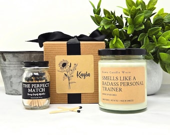 May include: A gift set featuring a candle, matches, and a decorative box. The candle has a label that reads "Smells Like a Badass Personal Trainer." The match jar is labeled "The Perfect Match." The box is adorned with a sunflower design and the name "Kayla."