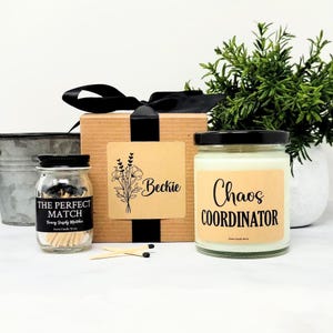 May include: A gift set featuring a brown cardboard box with a black ribbon, a glass jar of matches labeled THE PERFECT MATCH, and a glass candle jar labeled CHAOS COORDINATOR. The box has a label with a floral illustration and the name Beckie.