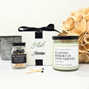 May include: A gift set with a white box tied with a black ribbon, a jar of matches, and a candle. The box has the name "Marissa" and a leaf design. The candle reads "In Loving Memory of Your Thirties."