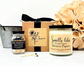 May include: A gift set featuring a square cardboard box with a black ribbon, a glass jar of matches labeled THE PERFECT MATCH, and a glass candle jar labeled Smells like Freshly Signed Divorce Papers, with a floral bouquet in the background.