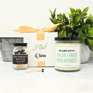 May include: A gift set featuring a white box with a peach ribbon, a small glass jar of matches labeled The Perfect Match, and a glass candle jar with a label reading The Calming Scent of a Color Coded Spreadsheet, all arranged together.