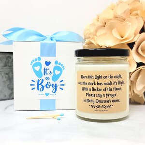 May include: A white gift box with a light blue ribbon and a label reading It's a Boy with blue footprints, alongside a glass candle jar with a black lid and a tan label featuring the text Burn this light on the night, once the stork has made it's flight. With a flicker of the flame, Please say a prayer in Baby Dawson's name. Two wooden matches lie in front.