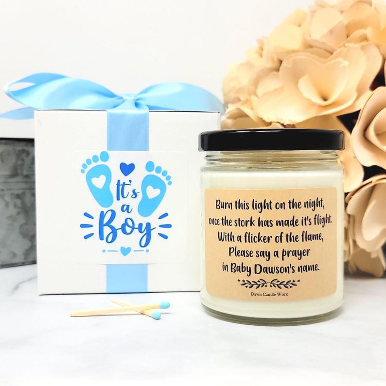 May include: A white gift box with a light blue ribbon and a label reading It's a Boy with blue footprints, alongside a glass candle jar with a black lid and a tan label featuring the text Burn this light on the night, once the stork has made it's flight. With a flicker of the flame, Please say a prayer in Baby Dawson's name. Two wooden matches lie in front.
