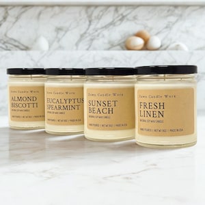 May include: Four clear glass jars filled with off-white soy wax, each topped with a black lid. The tan labels feature the text Dawn Candle Worx, the scent names Almond Biscotti, Eucalyptus Spearmint, Sunset Beach, and Fresh Linen, and Net Wt 9oz.