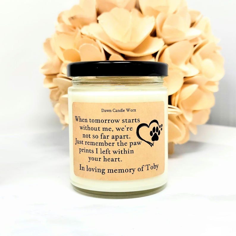 May include: A glass candle jar with a black lid and a cream-coloured wax fill. The label features the text Dawn Candle Worx and a poem about paw prints, accompanied by a heart and paw print graphic, dedicated to the memory of Toby.