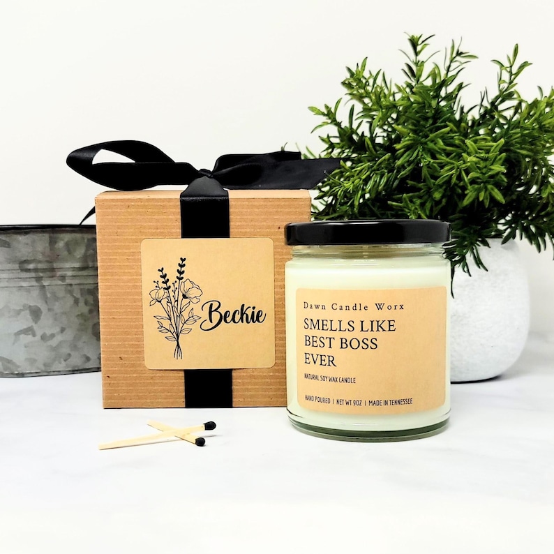 May include: A clear glass jar candle with a black lid and a tan label reading SMELLS LIKE BEST BOSS EVER, alongside a brown cardboard gift box with a black ribbon and a label featuring a floral illustration and the name Beckie.