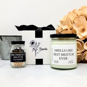 May include: A gift set featuring a white box with a black ribbon, a small glass jar of matches labeled THE PERFECT MATCH, and a glass candle jar with a white label reading SMELLS LIKE BEST MENTOR EVER, HAND POURED IN TENNESSEE, NET WT 9OZ.