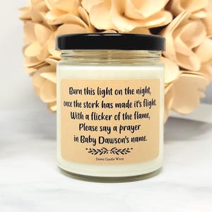 May include: A clear glass candle jar with a black lid containing white wax. A tan rectangular label features black text: Burn this light on the night, once the stork has made it's flight. With a flicker of the flame, Please say a prayer in Baby Dawson's name. Dawn Candle Worx.