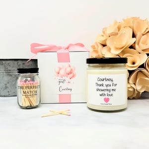 May include: A gift set featuring a white box with a pink ribbon, a jar of matches, and a candle. The box has a pink balloon graphic and the text "It's a girl! Courtney." The candle label reads "Courtney, Thank you for showering me with love."