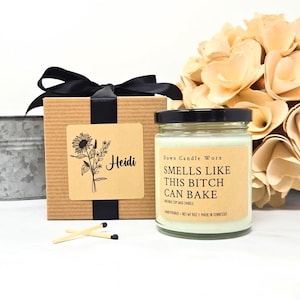 May include: A glass jar candle with a light-colored wax and a label reading "Dawn Candle Worx SMELLS LIKE THIS BITCH CAN BAKE" next to a brown gift box with a black ribbon, a sunflower illustration, the name "Heidi," and two wooden matches.