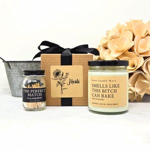 May include: A gift set with a candle, matches, and a decorative bouquet. The candle has a black lid and a label that reads "Smells Like This Bitch Can Bake." The match jar reads "The Perfect Match."