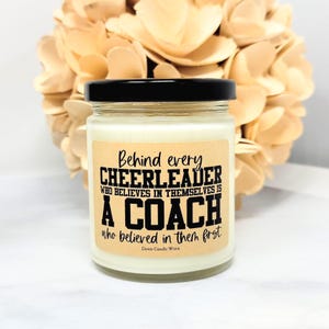 May include: A clear glass candle jar with a black lid and a light-colored wax fill. The label features the black text: Behind every CHEERLEADER WHO BELIEVES IN THEMSELVES IS A COACH who believed in them first. Dawn Candle Worx is printed below.