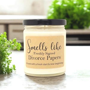 K&ouml;nnte beinhalten: Ein durchsichtiges Kerzenglas mit schwarzem Deckel, gef&uuml;llt mit cremefarbenem Wachs. Ein rechteckiges beiges Etikett auf der Vorderseite tr&auml;gt den schwarzen Text: Smells like Freshly Signed Divorce Papers, Infused with a fresh start & new beginnings.
