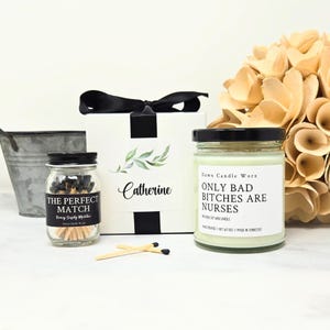 May include: A gift set featuring a white box with a black ribbon, a jar of matches labeled "THE PERFECT MATCH", and a candle with the text "ONLY BAD BITCHES ARE NURSES". The set includes a small metal bucket and dried flowers.