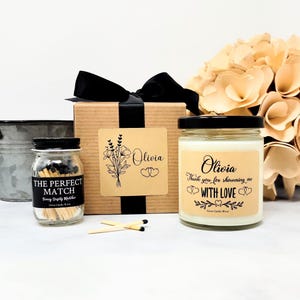 May include: A gift set featuring a brown cardboard box with a black ribbon, a small glass jar of matches labeled THE PERFECT MATCH, and a glass candle jar labeled Olivia Thank you for showering me WITH LOVE with floral and heart graphics.